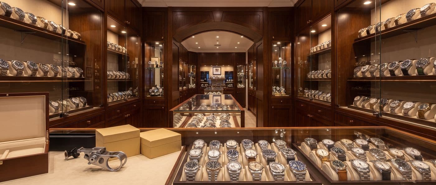 Independent pre-owned luxury watch dealer showroom, watches grouped by style in glass cases, an arched doorway to a back room, a loupe and watch boxes on the counter